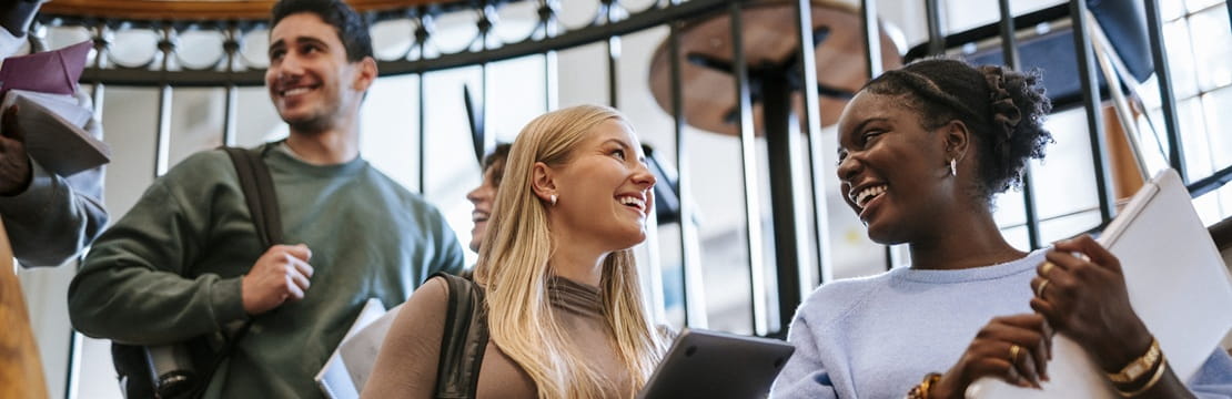 Students carrying backpacks and tablets walk through a campus building, representing the educational goals families can help support through a 529 college savings plan.